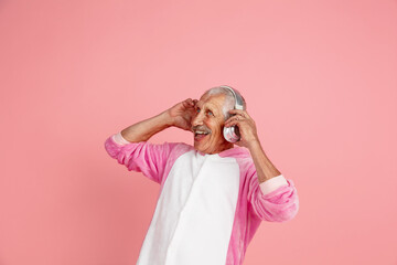 Laughting, dancing with headphones. Portrait of senior hipster man in stylish attire isolated on pink studio background. Tech and joyful elderly lifestyle concept. Trendy colors, forever youth © master1305