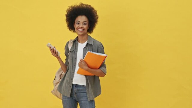 A positive woman is holding copy-books while using her smartphone standing isolated over a yellow background
