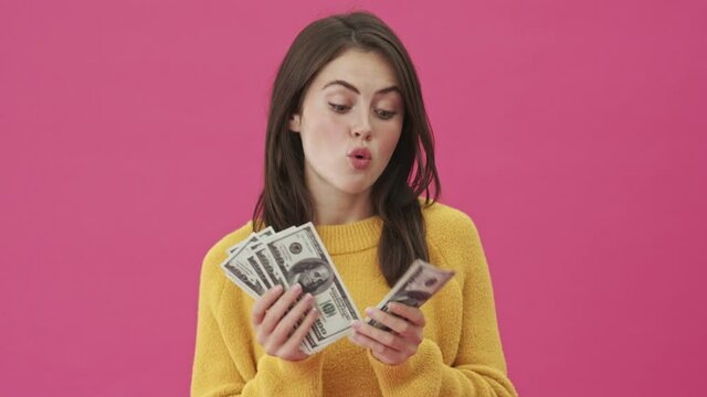 A pleased woman is counting her money standing isolated over a pink background