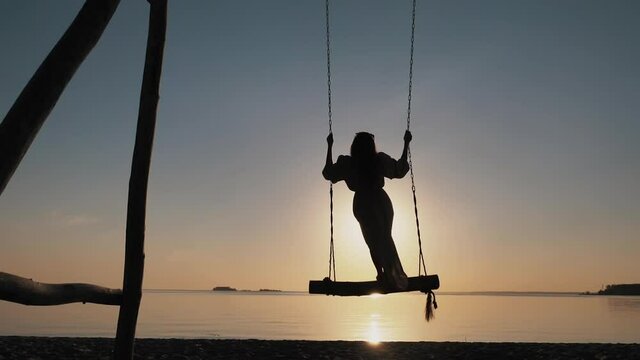Silhouette of young carefree girl swaying standing on wooden swings on amazing evening beach at sunset alone. Paradise sea relax, summer enjoyment, serene leisure, free happiness, recreation outdoors