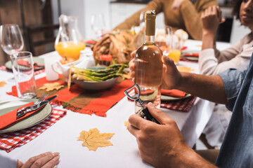 cropped view of man holding bottle of white wine and corkscrew during thanksgiving dinner with multicultural family