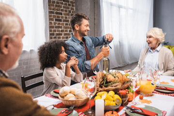 man opening bottle of white wine during thanksgiving dinner with multicultural family