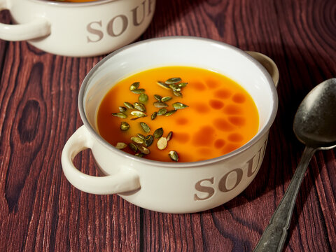 Two bowls of homemade pumpkin soup, decorated with pumpkin seeds and drops of pumpkin oil, on brown background