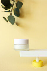 Mockup moisturizer cream in jar standing on abstract pedestal on pastel yellow background with copy space and defocused eucalyptus leaves, front view. Skincare beauty product template, vertical
