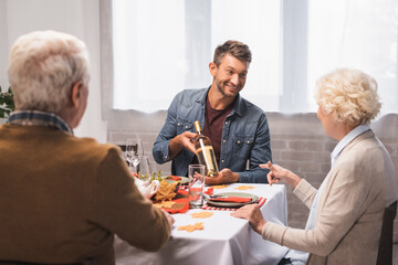 senior woman pointing with finger at wine bottle in hands of joyful son during thanksgiving dinner