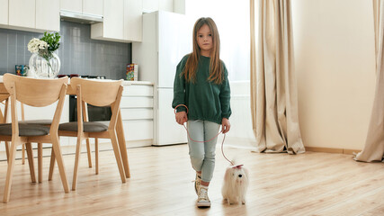 A cute little girl standing in a middle of a kitchen holding a leash of her white toy dog