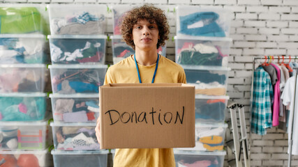 Caucasian guy holding donation box and looking at camera, posing in front of rack and boxes full of clothes, Young volunteer working for a charity