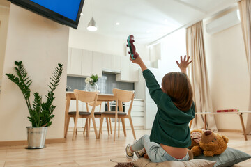 A small girl sitting on a floor in front of a TV cheering after winning a videogame holding a...
