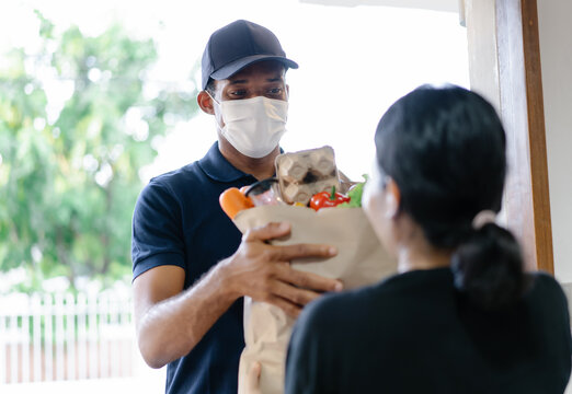 African American Delivery Man With Medical Mask Delivering Food To A Woman At Home.Online Shopping And Express Delivery. Under Quarantine, Disease Outbreak, Coronavirus Covid-19 Pandemic.