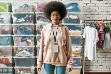 African american woman looking at camera, posing in front of boxes full of clothes, Young volunteer working for a charity