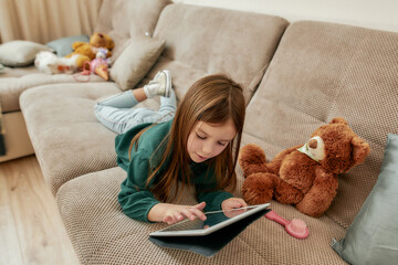 A small cute girl playing games on her tablet lying on her stomach on a sofa with her gumshoes on...