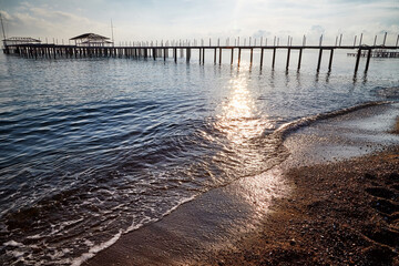 View from sand beach to water of sea, waves and pier in a nice day or evening with blues sky, bright sun and white clouds. The concept of a holiday on the sea or ocean in the South.