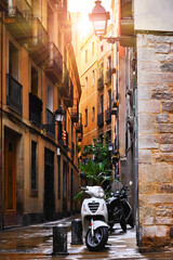 Barcelona, Catalonia, Spain. Romantic narrow residential bystreet in Gothic Quarter. Antique street lamp at wall of old house, parked motorbikes and arch with window between buildings.