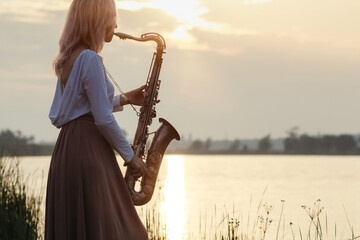 silhouette of a young beautiful girl playing the saxophone at sunrise by the river, a woman in a long dress on the nature at sunrise relaxing, concept music - obrazy, fototapety, plakaty