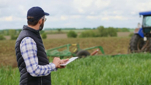 Bearded farmer wearing a cap uses a specialized app on a digital tablet PC on the background of working tractor with cultivator in the field