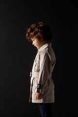 Profile of fashionable adorable curly little boy, wear in trench, poses with his head down, over black background.