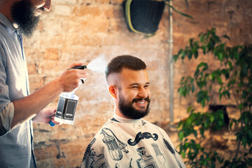 Hairdresser using hair spray on client hair at barber shop. Good looking man visiting barber shop. 