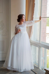 Full-length portrait of the bride in a white wedding dress by the window
