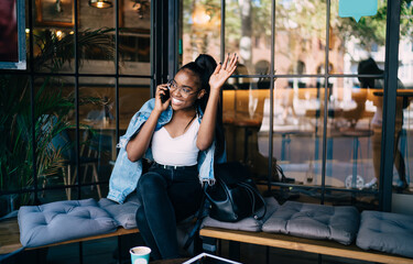 Smiling african american woman in casual wear sitting in cafe interior talking on mobile phone and waving to somebody, cheerful dark skinned female satisfied with positive smartphone conversation