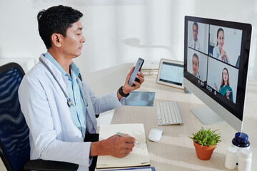 Serious middle-aged Asian doctor having online conference with colleagues and taking notes in planner