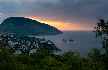 Sea bay with rocks "Adalary" sticking out of the sea. Town on the shore. Morning, calm sea, from behind Mount Ayu-Dag (Bear Mountain) the rays of the rising sun. Gurzuf, Yalta, Crimea.