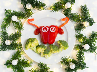 Funny plate with symbol of 2021 bull cow made of vegetables: sweet pepper, broccoli, tomato and onion with pine branches and balls. Vegan or vegetarian food art for New Year or Christmas celebration