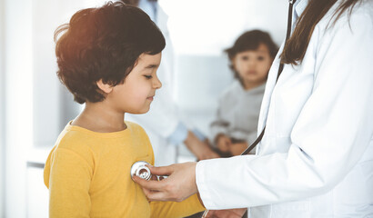 Obraz premium Woman-doctor examining a child patient by stethoscope in sunny clinik. Cute arab boy at physician appointment