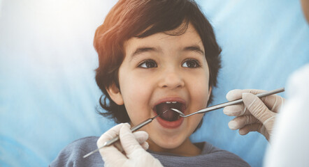 Cute arab boy sitting at dental chair with open mouth during oral checking up with doctor. Visiting sunny dentist office