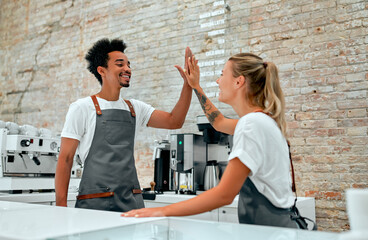 Caucasian female barista and African attractive male barista making high five gesture behind the counter in a coffee shop.