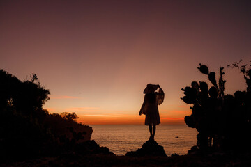 Silhouette woman in straw hat standing on top of a cliff near cactus and look at the sea at sunset