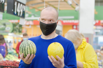 Fototapeta premium Male in a black medical mask stands in a supermarket. He has a melon and a watermelon in his hands, the man can choose what to buy. Background, you can see products and people in protective masks.