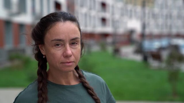 serious middle-aged brunette woman is looking at camera, standing in yard of apartment building at summer day