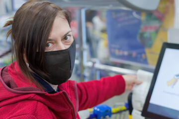 Shop, girl is wearing a medical mask. Girl pays for purchases and takes a cash receipt. She looks at the camera. Girl is wearing a red jacket. Products payment terminal is visible in the background.