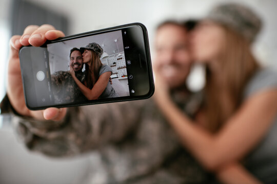 Young soldier taking selfie with his wife at home. Loving couple sitting on sofa in living room..