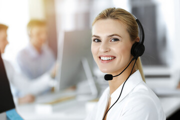 Blond business woman using headset for communication and consulting people at sunny office. Call...