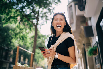 Overjoyed beautiful asian female looking at camera laughing at funny content from social networks,cheerful woman amazed with good news using smartphone connected to 4G on sunny day in town