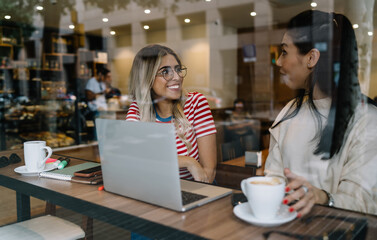 Positive multiracial female best friends communicating enjoying positive conversation on meeting, smiling young 20s women colleagues spending break talking to each other sitting in cafe interior