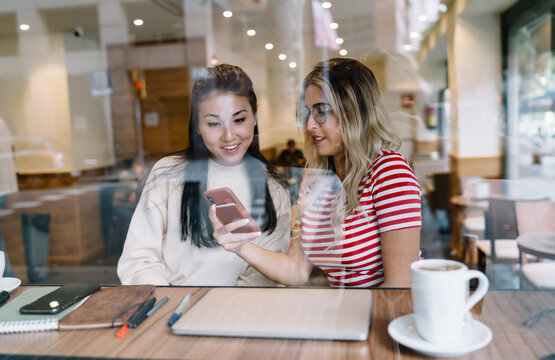 Smiling multiracial female friends sitting in cafeteria on free time watching funny video on smartphone together, positive caucasian girl showing photo on mobile phone to asian best friend on meeting
