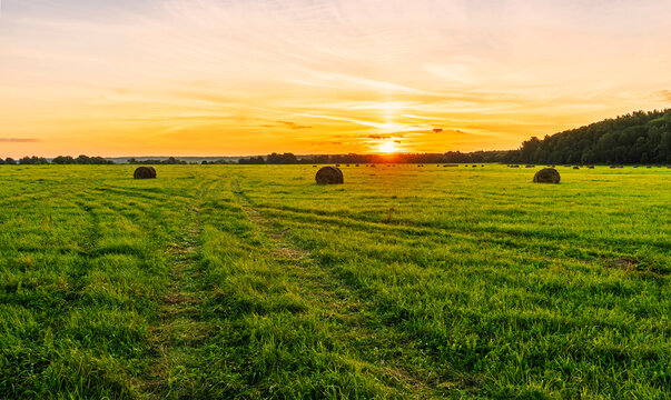 Scenic view at beautiful sunset in green shiny field with hay stacks, bright cloudy sky, country road and golden sun rays with glow, summer valley landscape © Yaroslav