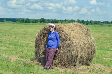 Naklejka na ściany i meble Rural area, next to a haystack stands a happy girl in a blue jacket and sunglasses. Summer, the sun is shining bright and white clouds are running across the blue sky.
