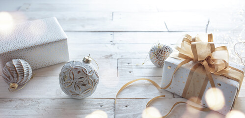 Gift with golden ribbon bow and christmas baubles on white wooden vintage background. Horizontal close-up with festive bokeh and short depth of field for a christmas concept.