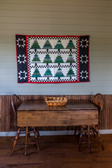 A country rural style quilt tapestry hanging on the wall above a small antique wood table with two chairs as a small dining room inside a rustic hunting and fishing cabin 