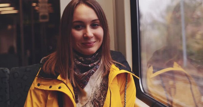 Happy Young Woman Looking Out of a Train Window. SLOW MOTION 4K. Relaxed smiling girl seating near the window in city train during her daily commute. Enjoying life.