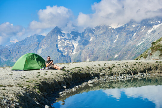 Male tourist sitting in chair near tent in the rocky alpine mountains near lake with fresh clear water and using smartphone in sunny warm summer day. Concept of camping, travelling, nature, gadgets.