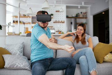 Smiling young man using VR headset at home on couch. Woman and her husband enjoying virtual reality in apartment..