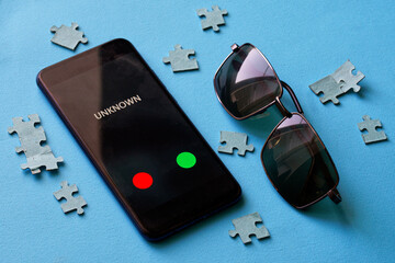 A smartphone with an unknown call lies on a blue surface next to a puzzle and sunglasses. The...