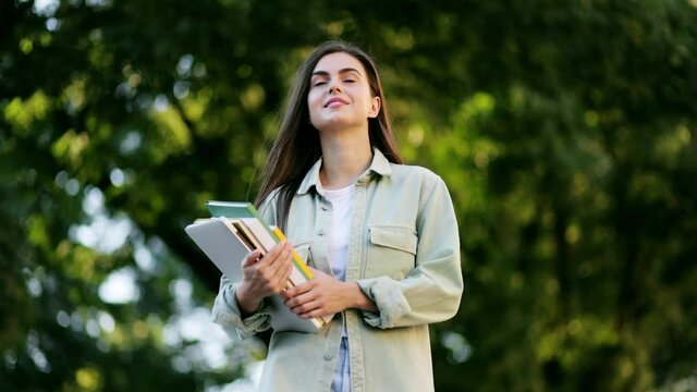 Happy young beautiful female student walking in campus park. Portrait of charming young lady holding books.