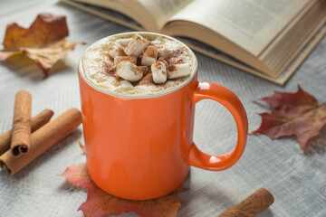 Coffee with whipped cream and marshmallow in bright orange mug on grey table. An open book in soft focus in background. Fall time.