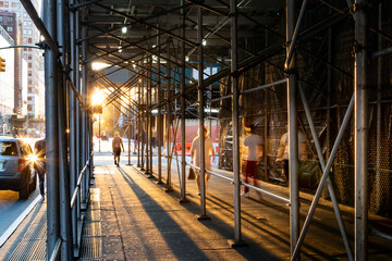 People walking under construction scaffolding on the streets of New York City with the light of sunset shining in the background