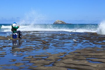 Naklejka premium Man fishing from a rocky shore, with waves breaking behind him. Muriwai Beach, New Zealand